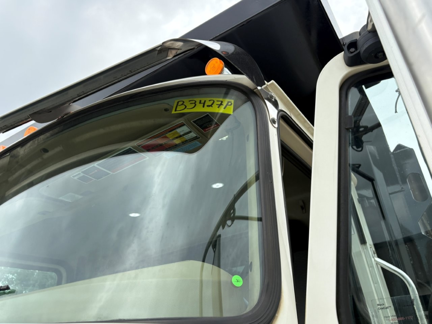 Truck cab window with yellow tag reading B3427P on upper windshield. Partial view of Mack GR64F dump truck, beige cab, open door.