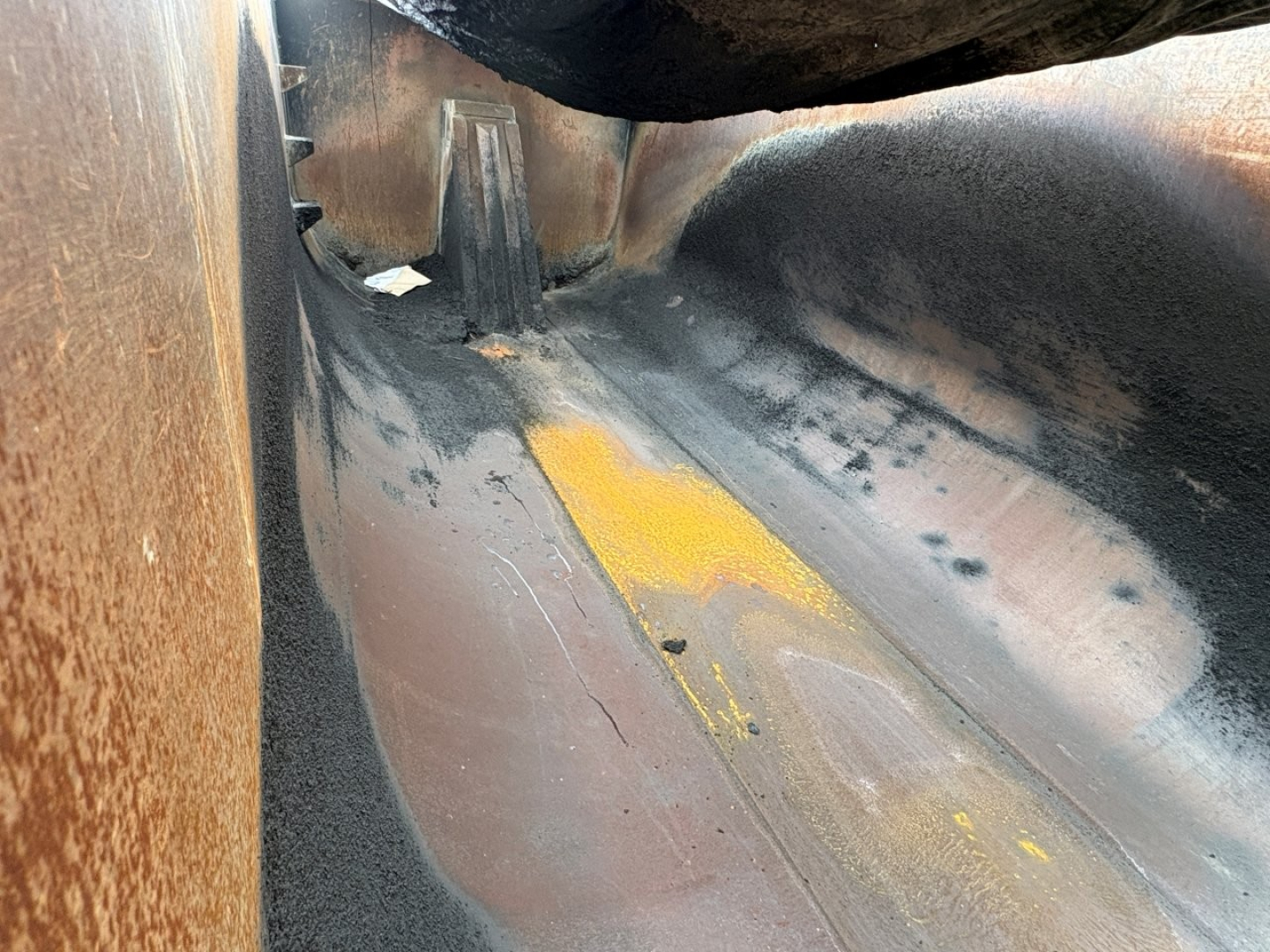 Dump truck bed interior showing rust-colored metal and yellow paint marks; fuel or material residue on curved surface.