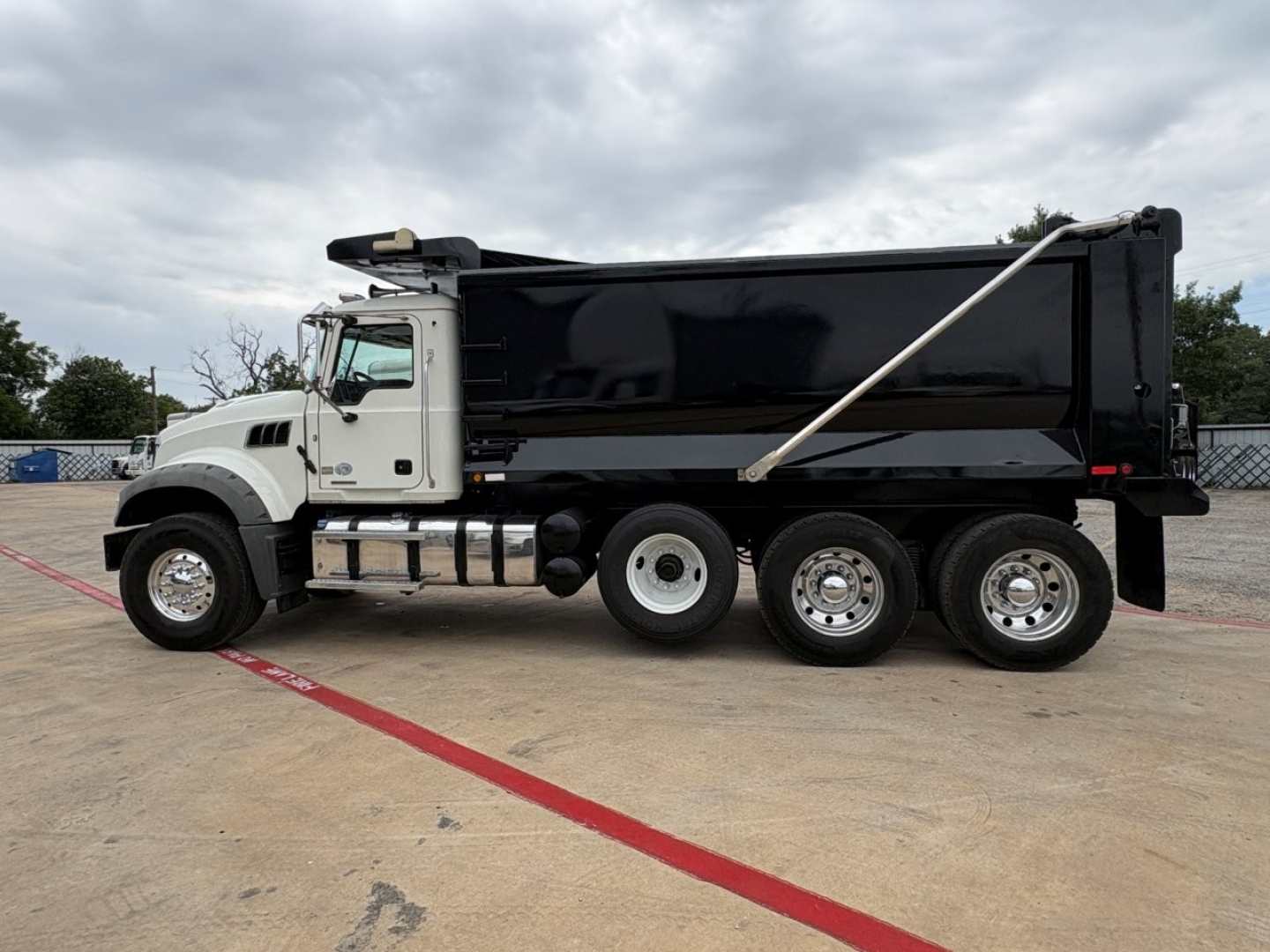 Dump truck side view showing Mack GR64F dump bed with black sides, white cab, three rear axles, chrome wheels, and hydraulic hoist. No visible text legible on the truck body.