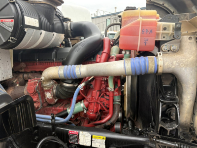 Engine bay of 2020 Mack GR64F dump truck showing red engine block, blue hoses, coolant reservoir, and air intake; visible VIN tag and assorted components.