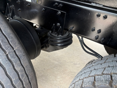 Truck frame undercarriage with suspension and axle components visible; drum-like bellows and braided hoses; front wheel and tire in view.