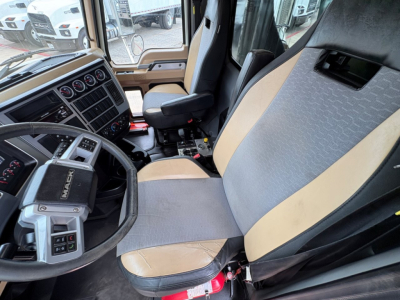 Truck cockpit showing Mack GR64F driver's seat with beige and gray upholstery, steering wheel, dashboard gauges, and center console.