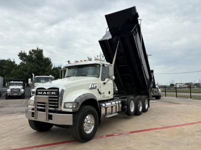 2010 Mack GR64F dump truck with raised dumping bed in white cab and black bed, front grille with Mack logo, side fuel tanks, heavy-duty tires.