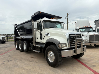 Dump truck front-right view showing Mack GR64F dump body, white cab, chrome grille, dual rear wheels, and black dump bed.