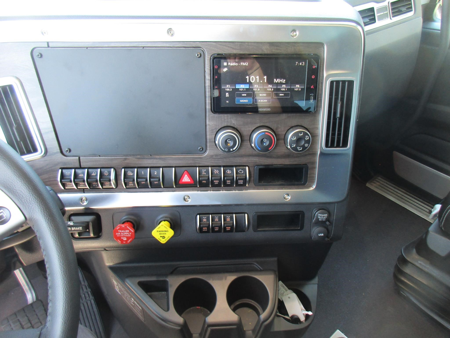 Dashboard of a 2027 Western Star 47X showing a digital infotainment screen at center, climate controls, multiple switches, cup holders, and a yellow and red hazard label cup. Visible text includes 101.1 MHz on the radio, various control lab