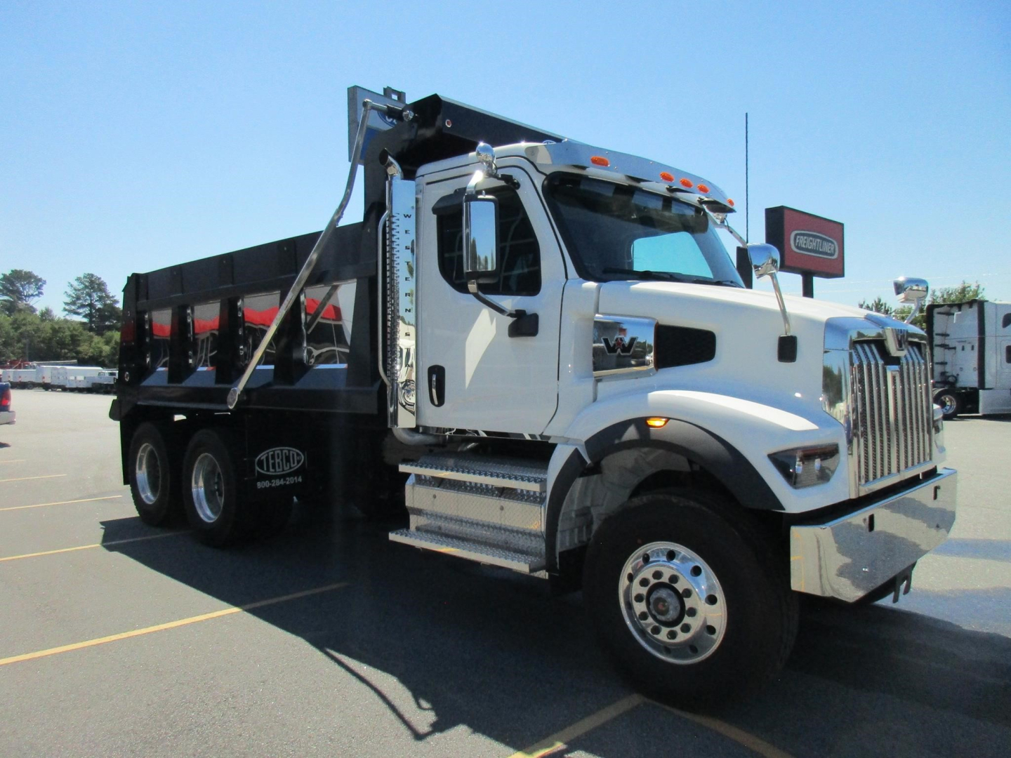 White Western Star dump truck with black dump body in a parking lot. Front grille with chrome, large side mirrors, and two-axle front with chrome steps. Text on cabin and wheels not legible.