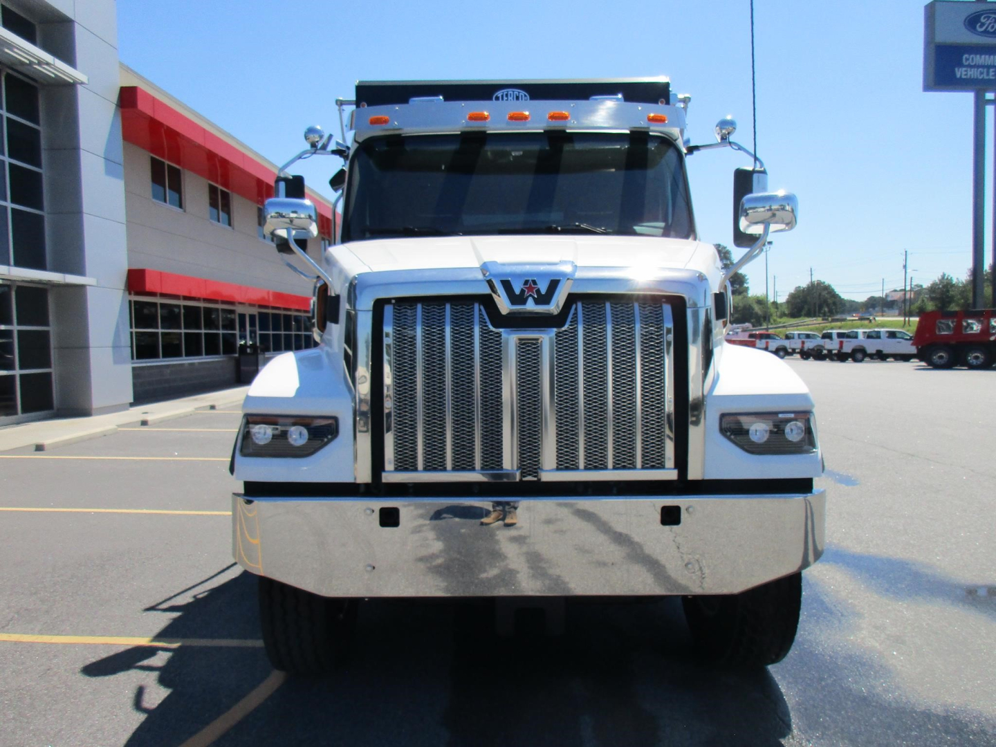 Front view of a white Western Star 47X dump truck in a dealership lot, chrome grille, emblem centered, large bumper, side mirrors, and building in background.