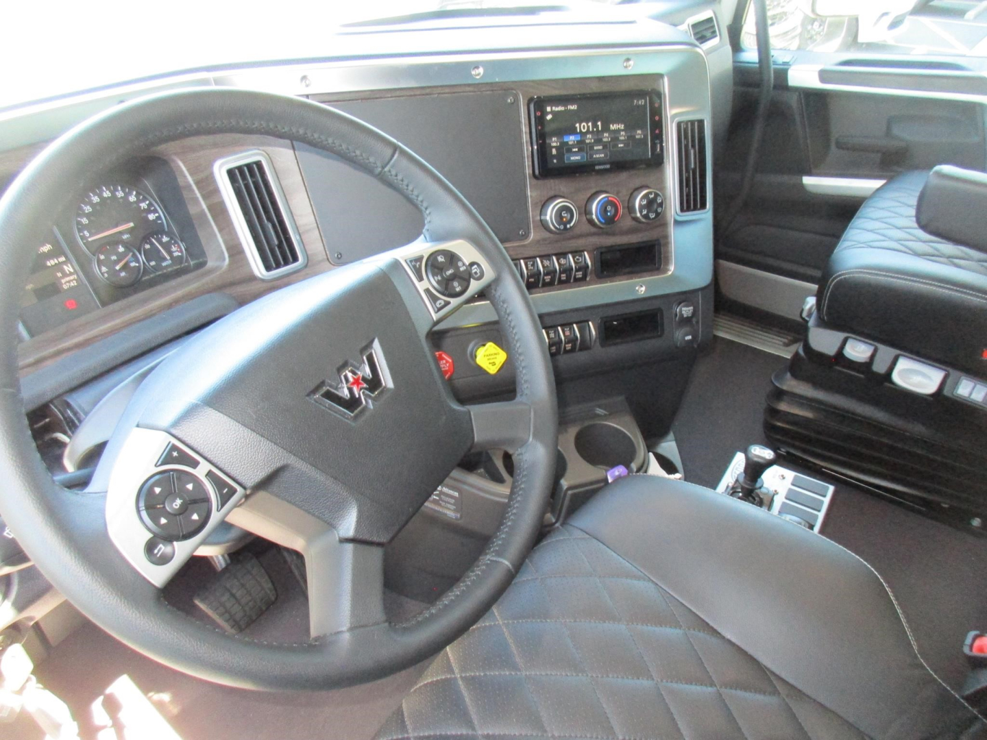 Interior of 2027 Western Star 47X dump truck showing steering wheel with logo, dashboard display, center console, gear selector, and quilted black seat.