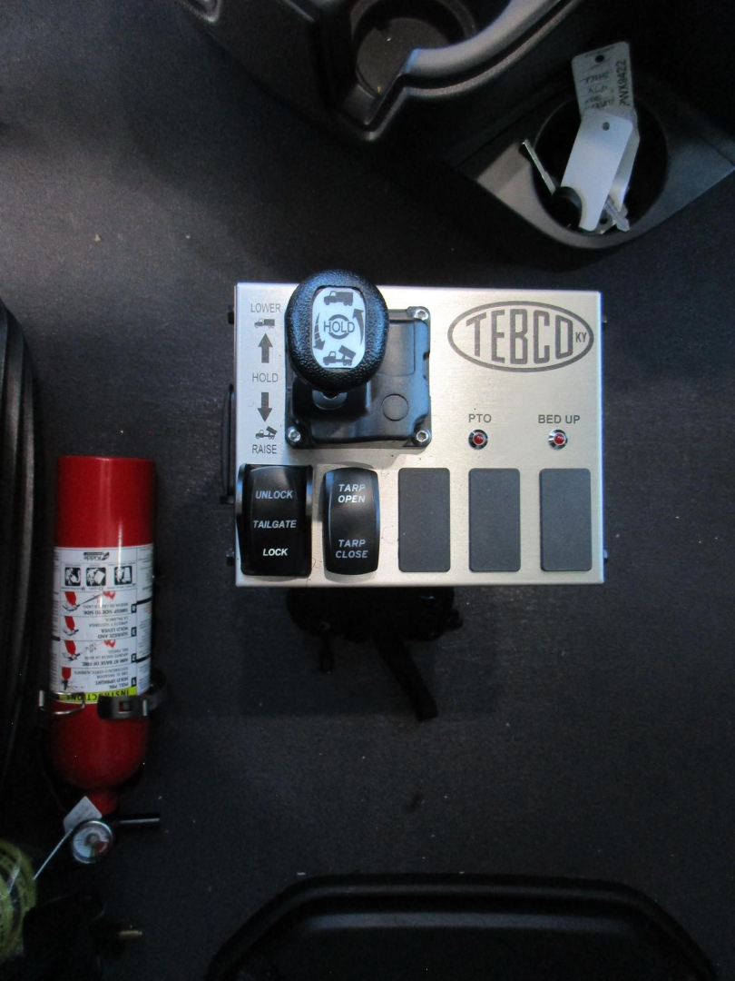 Panel with TEBCD controller and ignition key on a truck dashboard area; fire extinguisher visible on left; labeled switches and grip, plus cylindrical fuse/valve near.
