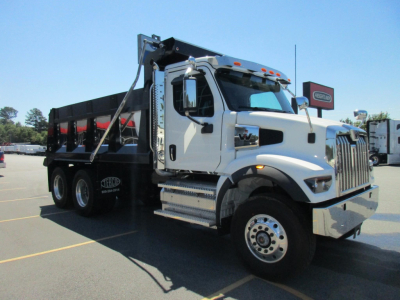White Western Star dump truck with black dump body in a parking lot. Front grille with chrome, large side mirrors, and two-axle front with chrome steps. Text on cabin and wheels not legible.