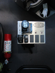 Panel with TEBCD controller and ignition key on a truck dashboard area; fire extinguisher visible on left; labeled switches and grip, plus cylindrical fuse/valve near.