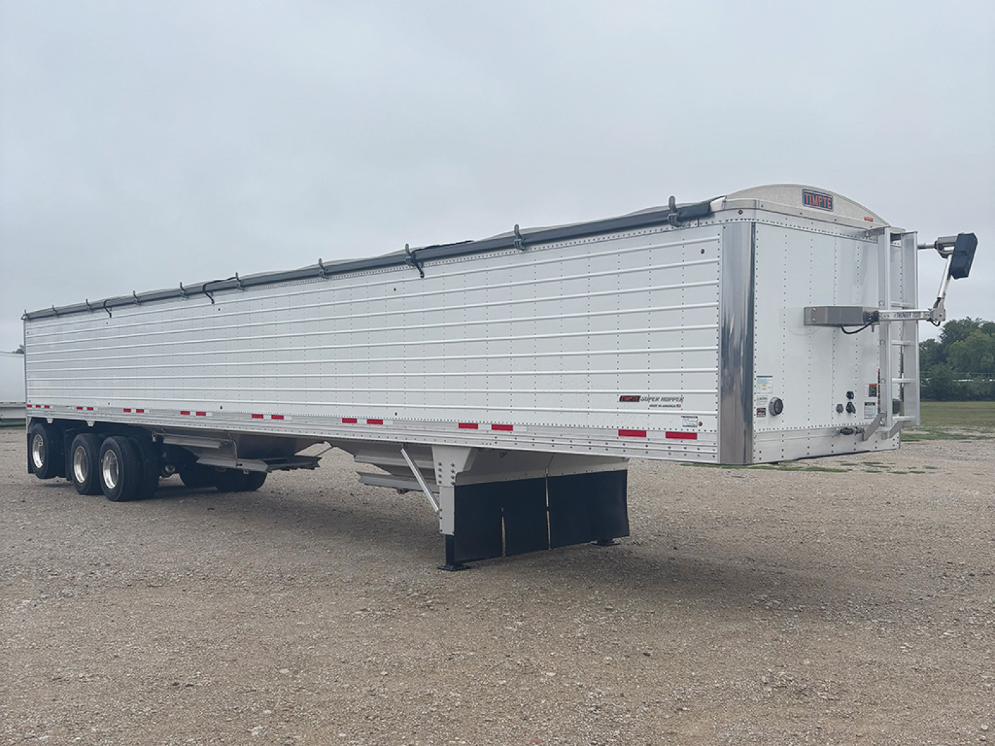 Timpte Super Hopper trailer side view showing long aluminum body, multiple axles, and a front landing gear jack. Visible markings on rear ramp area.