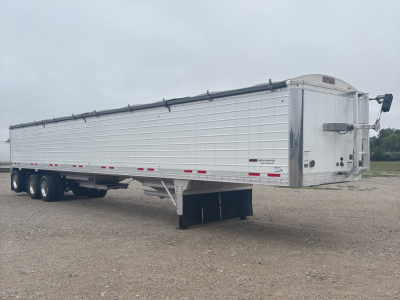 Timpte Super Hopper trailer side view showing long aluminum body, multiple axles, and a front landing gear jack. Visible markings on rear ramp area.