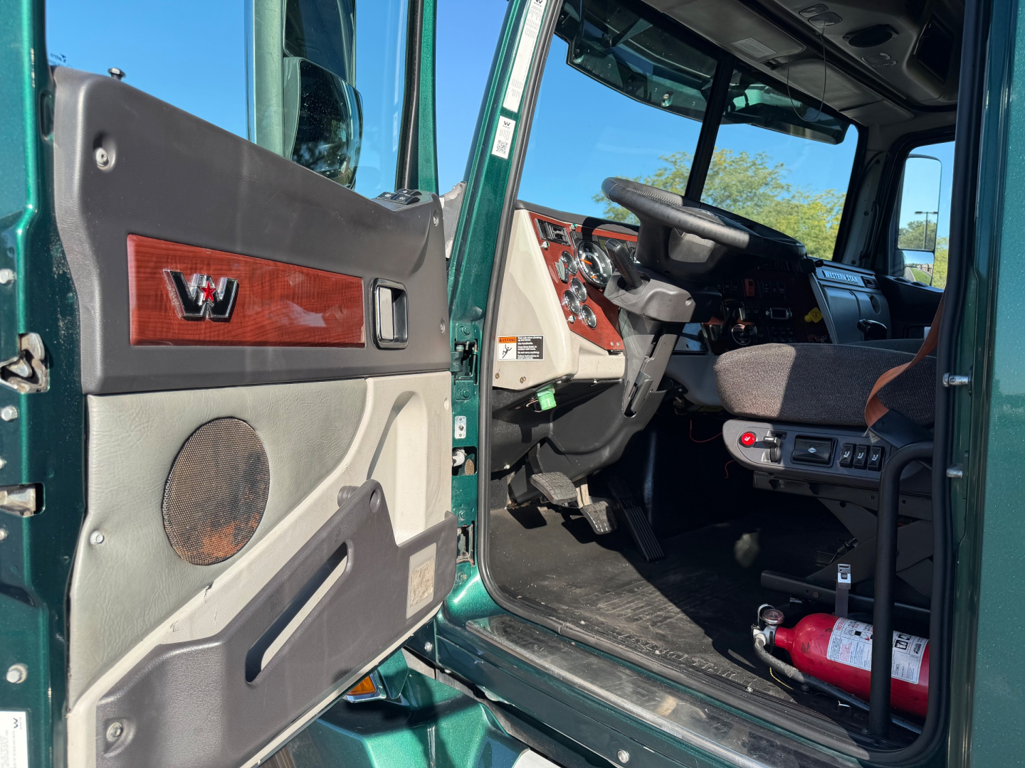 Cab interior of a 2017 Western Star 5700XE truck showing green exterior, driver door open, steering wheel, dashboard, and red fire extinguisher in the cabin.