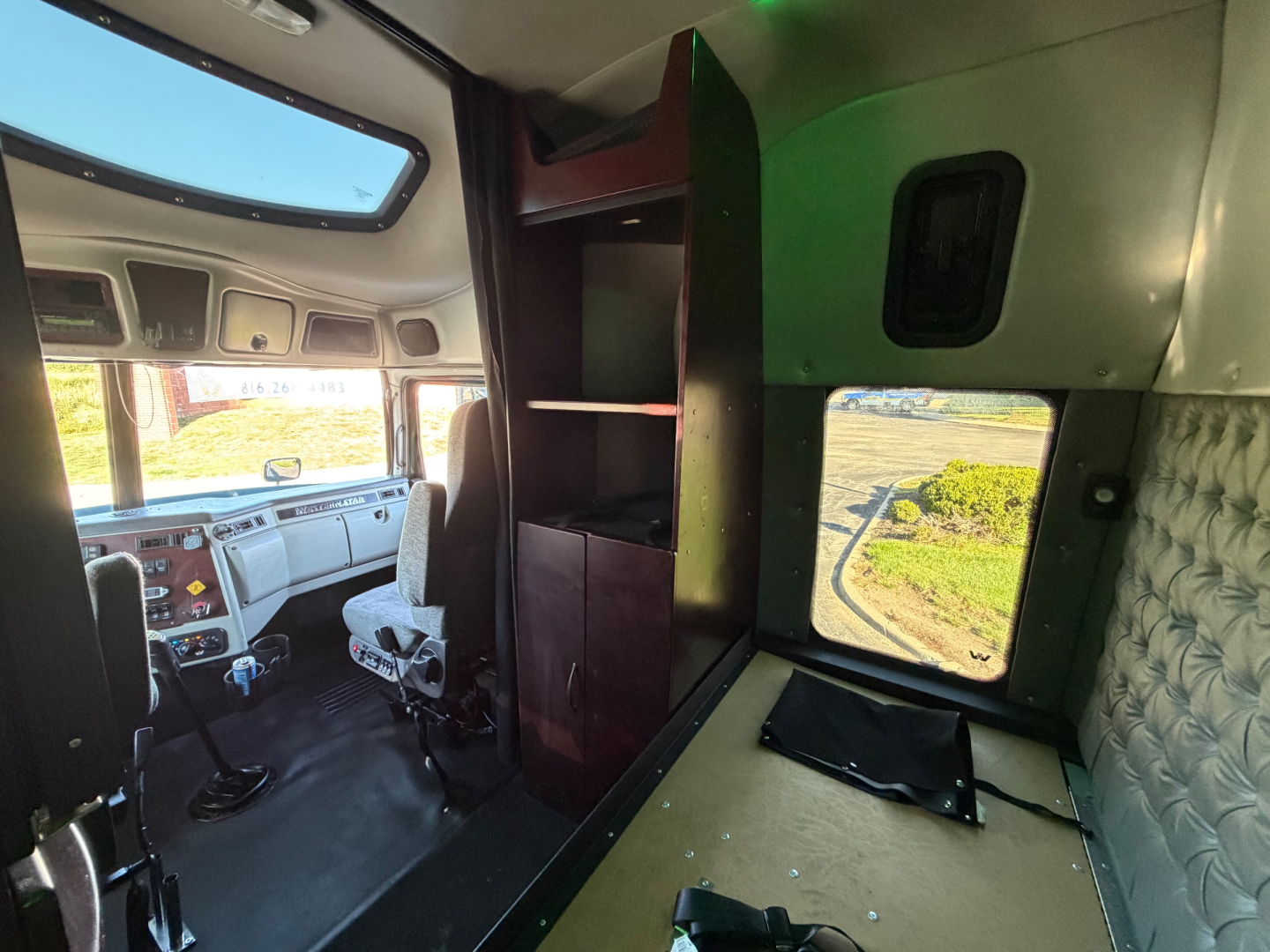 Truck cab interior showing sleeper area with overhead storage, window, and seating area; dashboard and steering visible at left; green wall panel and fabric quilted side wall.