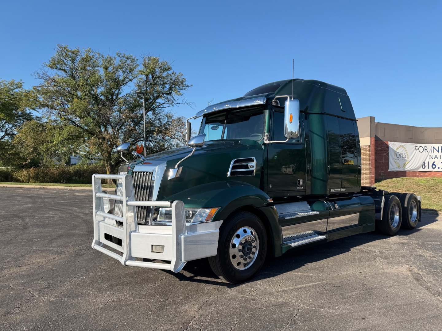 Green 2017 Western Star 5700XE tractor in a parking lot, front grille guard visible, sleeper cab, chrome wheels, three-axle tractor, sunlight clear sky.