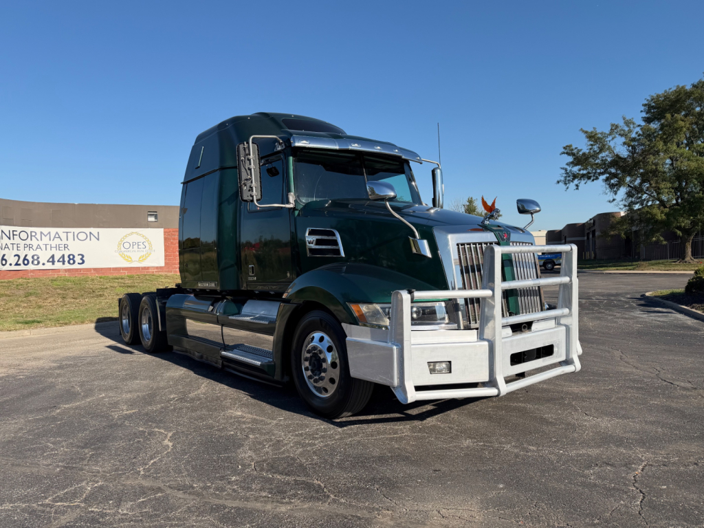 Green Western Star 5700XE tractor-trailer in a parking lot with chrome bumper guard and dual rear axles. No visible text on truck; sign in background reads 'INFORMATION 972-666-4483' but not on vehicle.
