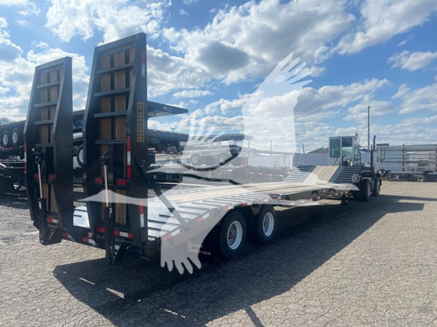 Trailer showing low-profile drop deck trailer with dual ramps and six wheels on the side, view from rear-left, blue sky and parking lot.