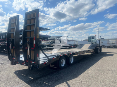 Drop deck trailer showing tall loading ramps raised, tandem axles, and long flatbed with steel decking.