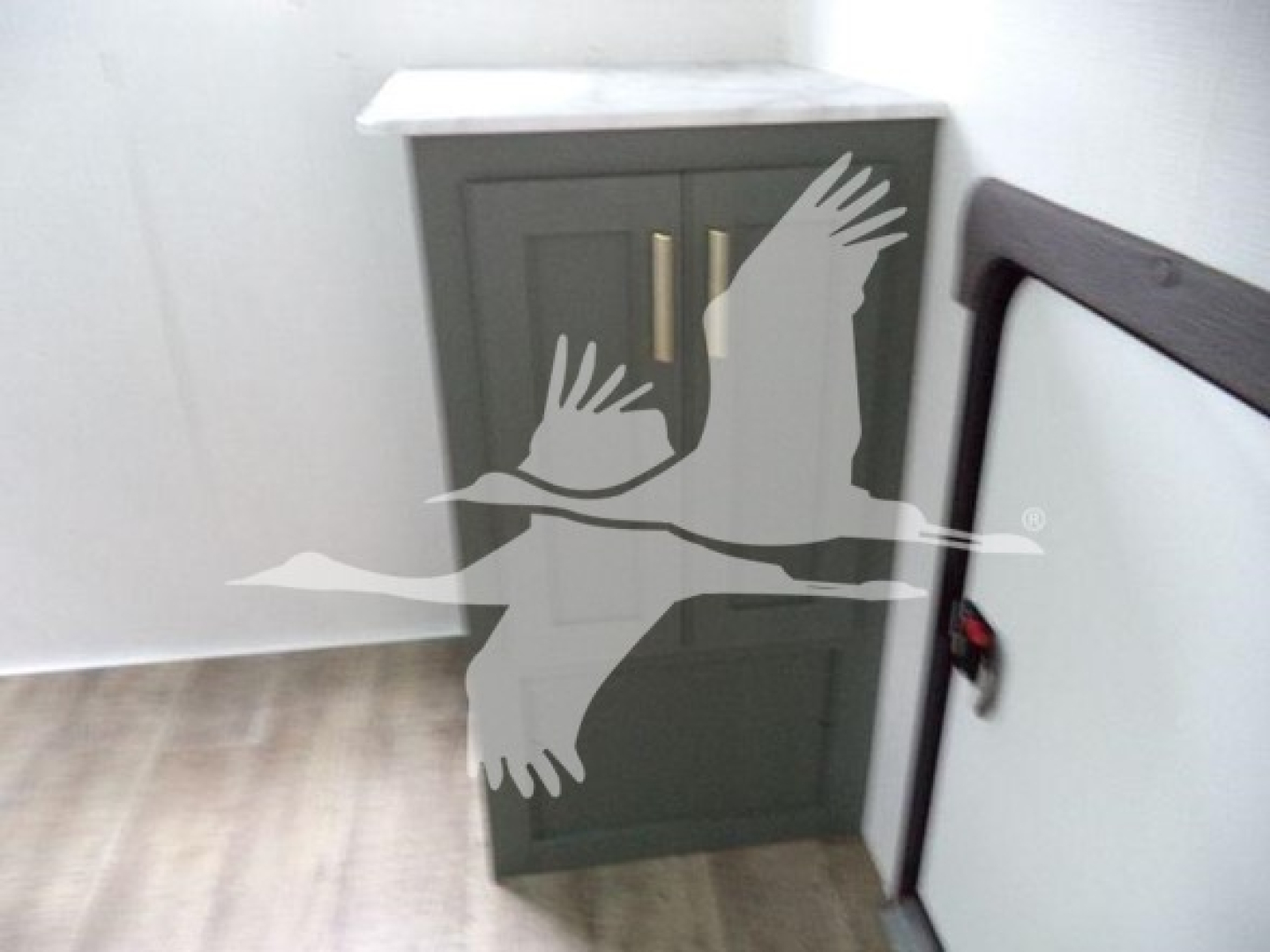 Trailer interior showing a gray cabinet with gold handles and a white countertop, with a white wall and light wood floor.