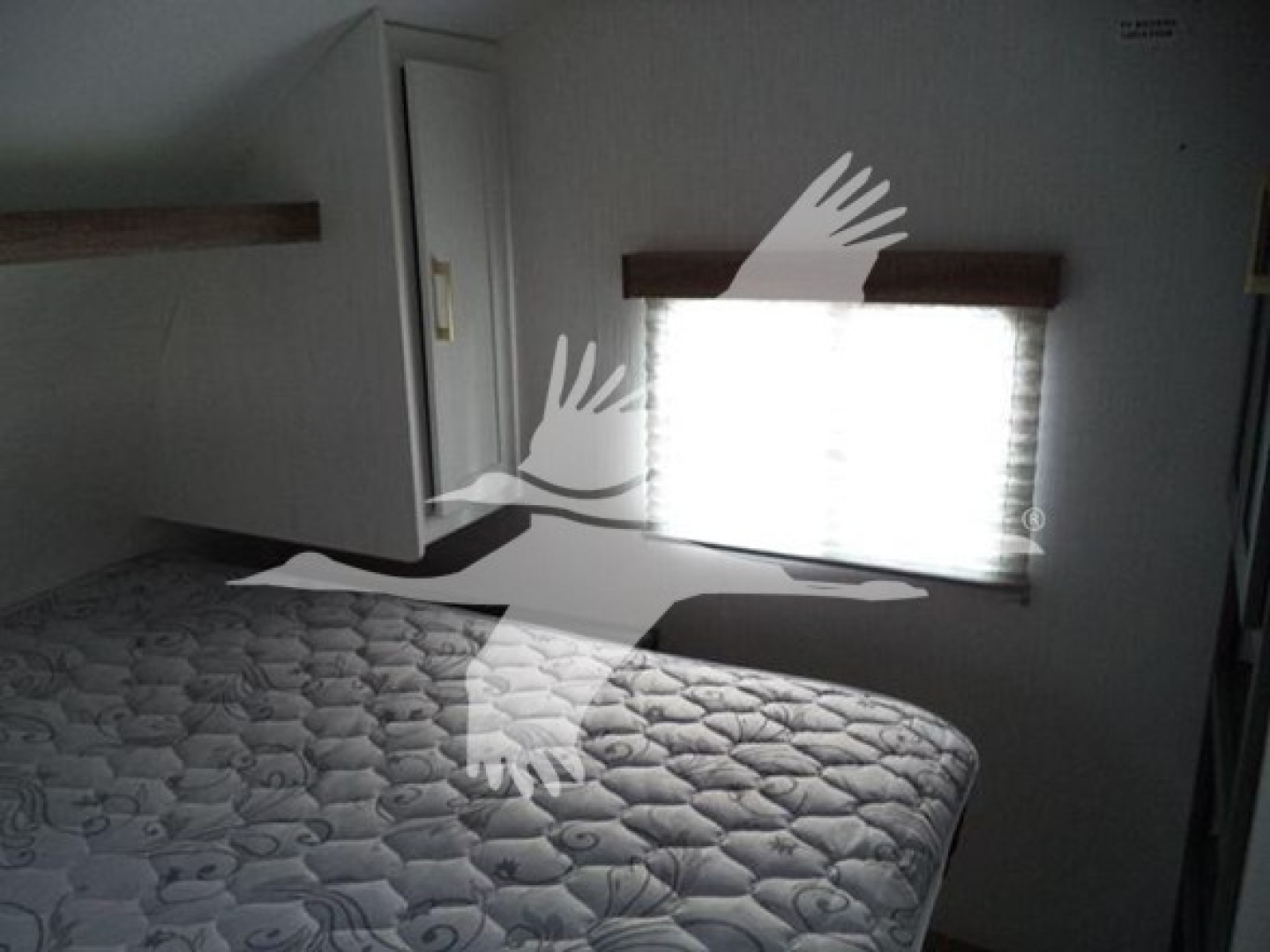 Travel trailer interior showing a bed with quilted mattress and a window with shade. No clear labels or fluid reservoirs visible.