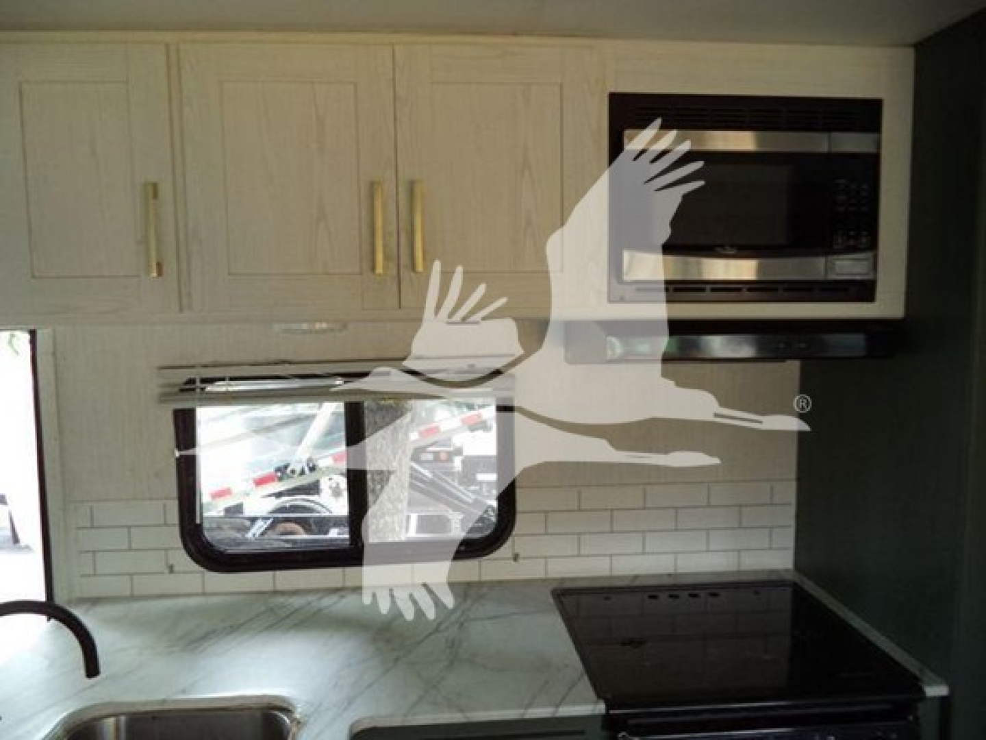 Interior of a trailer kitchen showing upper cabinets, microwave, window, stove, and sink area in a compact layout.
