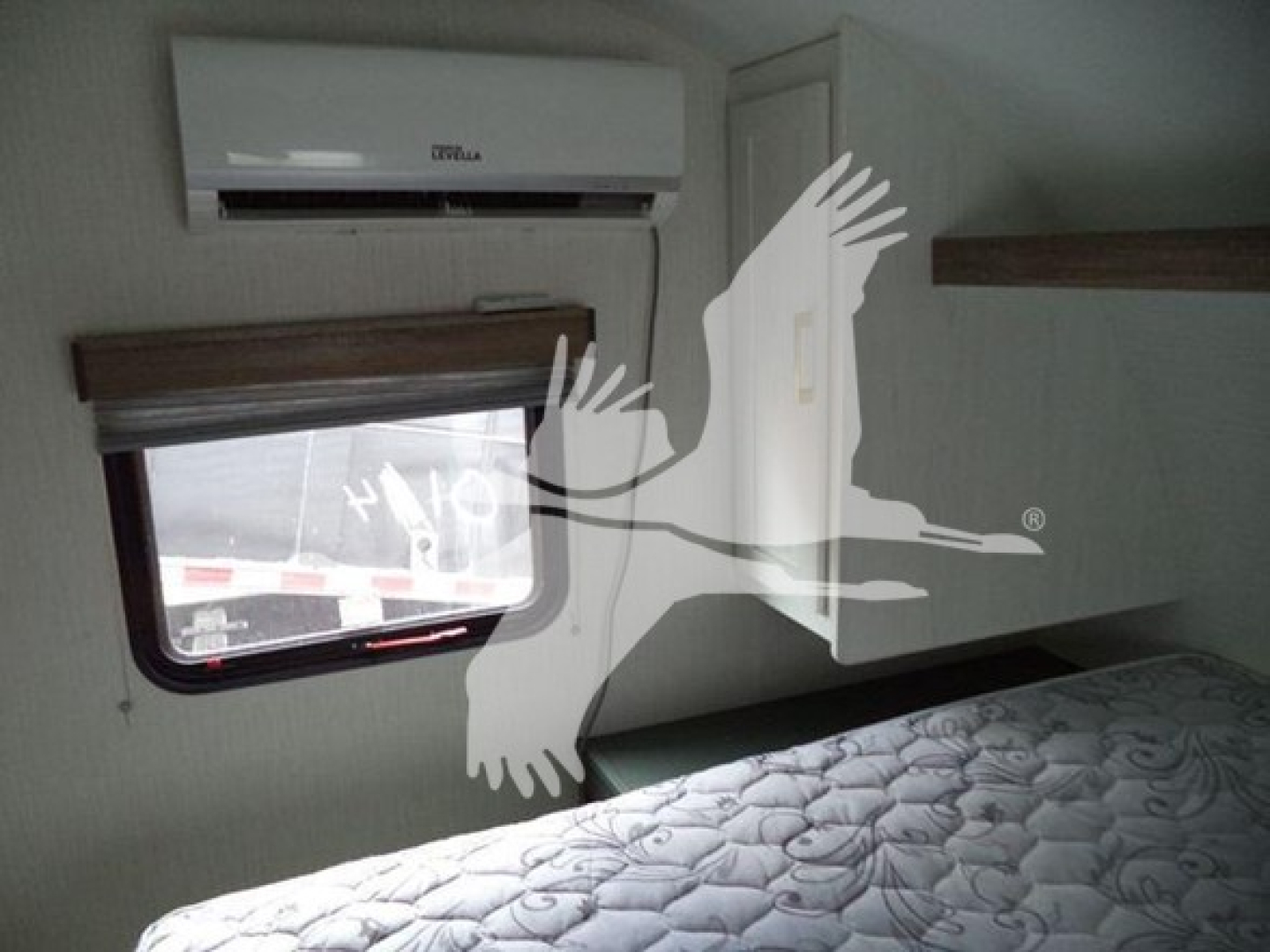 Trailer interior showing bed, window with shade, air conditioner unit, and overhead cabinet.
