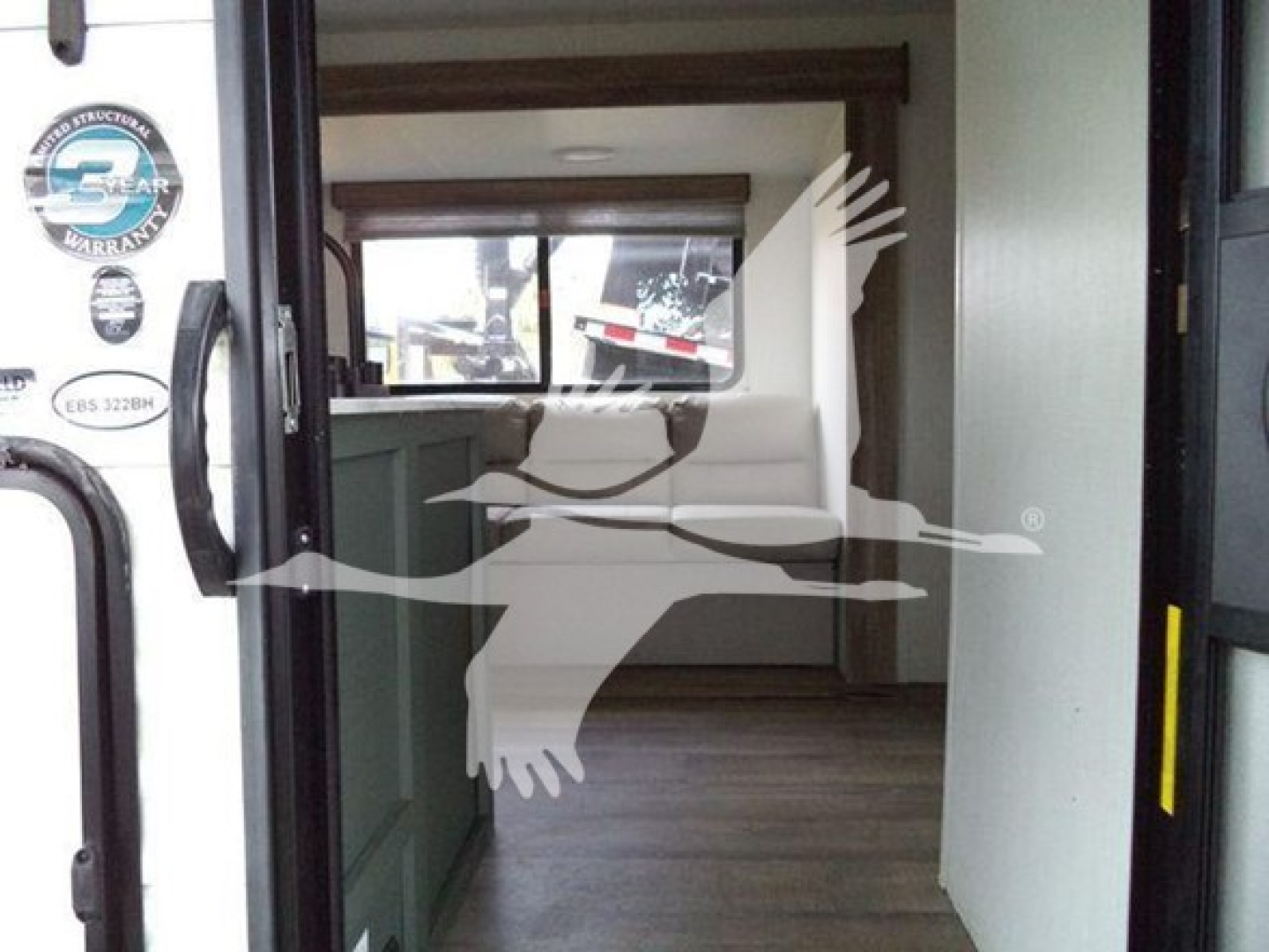 Travel trailer interior through open doorway, kitchenette on left, seating area and window at far end.