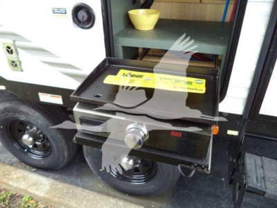 Exterior view of a travel trailer hitch and drop-down scissor storage box mounted on a towable trailer with a spare wheel nearby. Visible text includes a yellow label on the folded compartment with the brand 'LATCH' or similar and safety wa
