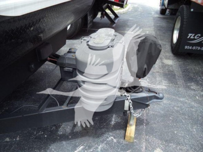 Trailer hitch with covered propane tank and coupler, on a trailer frame; chain and wooden block support visible; vehicle wheel and fender in background.