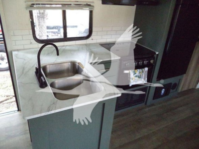 Kitchen area in a travel trailer showing a countertop with a sink and faucet, a window above, and a stove/oven unit to the right.