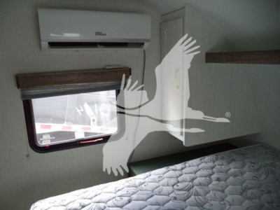 Trailer interior showing bed, window with shade, air conditioner unit, and overhead cabinet.