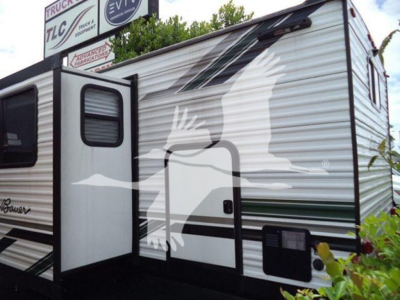 Travel trailer with graphic bird decal on white aluminum side, open entry door, slide-out visible, RV exterior with green stripe accents.