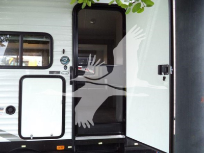Travel trailer entry door open showing interior. Exterior has white aluminum panels with black framed window and a side hatch with black frame.