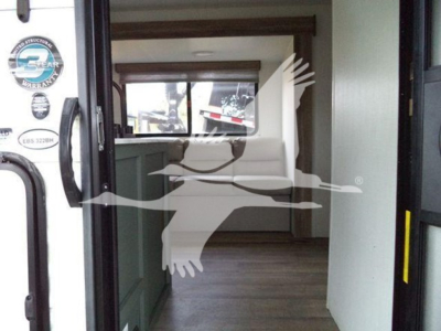 Travel trailer interior through open doorway, kitchenette on left, seating area and window at far end.