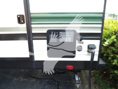Rear of travel trailer shows exterior access panel, electrical inlet, and stabilizer hitch with electric cord plugged in.