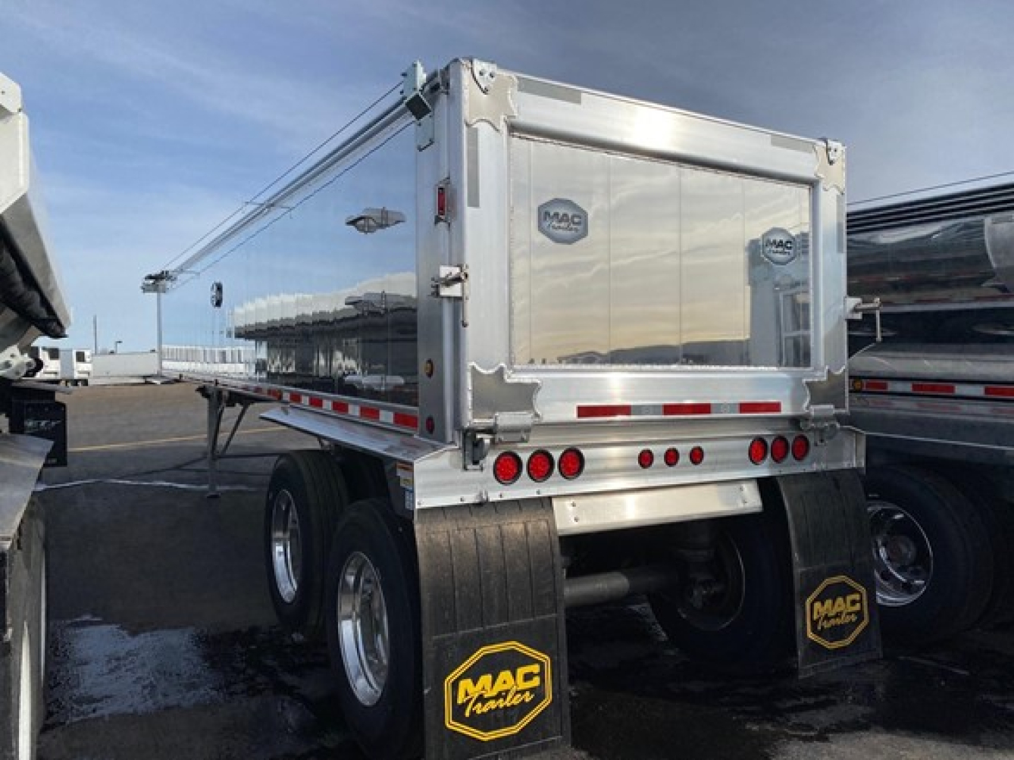 Dump trailer in side view showing polished aluminum walls, rear MAC logo, taillights, and dual rear axles with mud flaps.