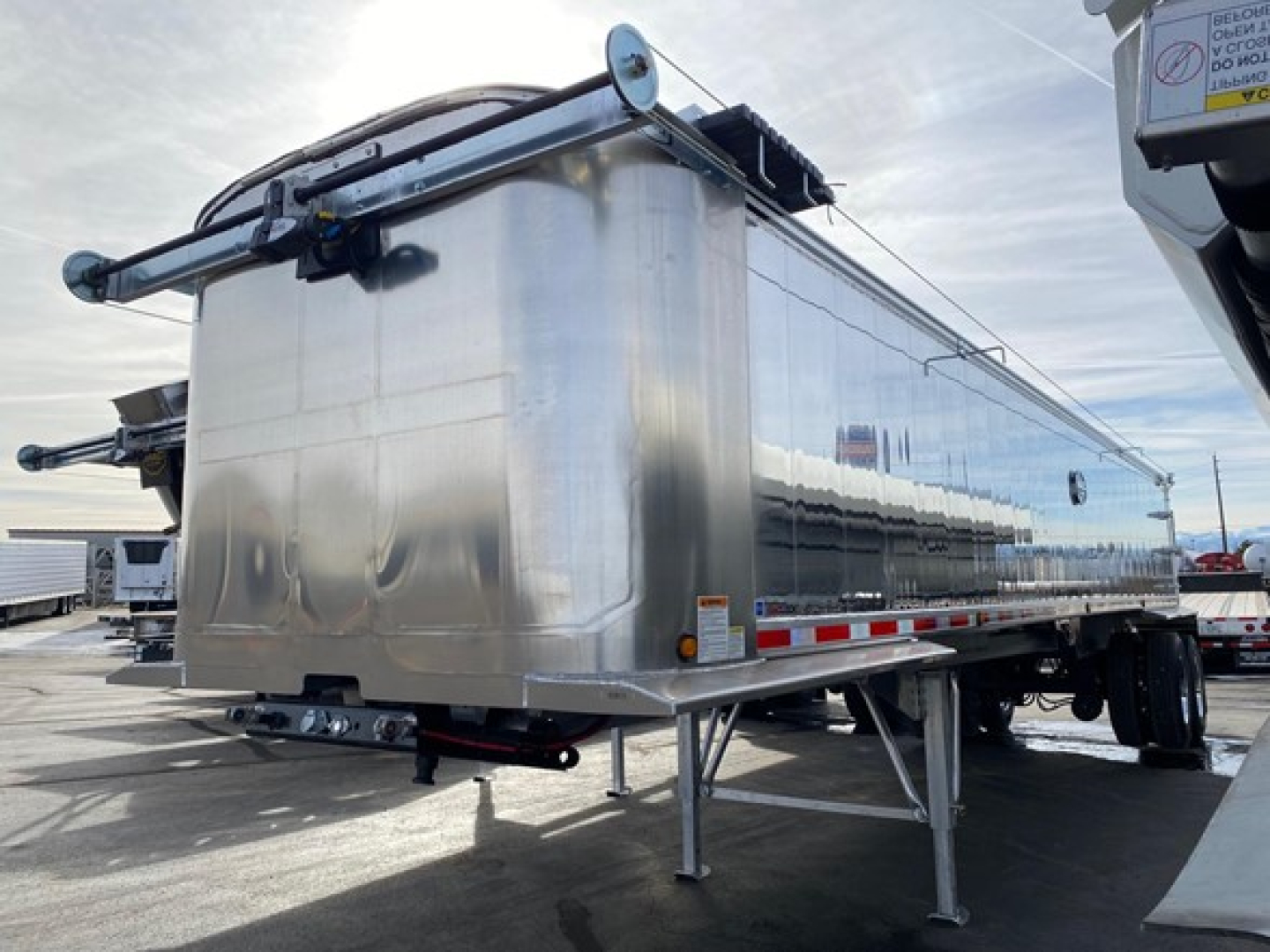 Dump trailer by Mac Western with polished aluminum body and rear door, front landing gear visible, blue sky reflection.