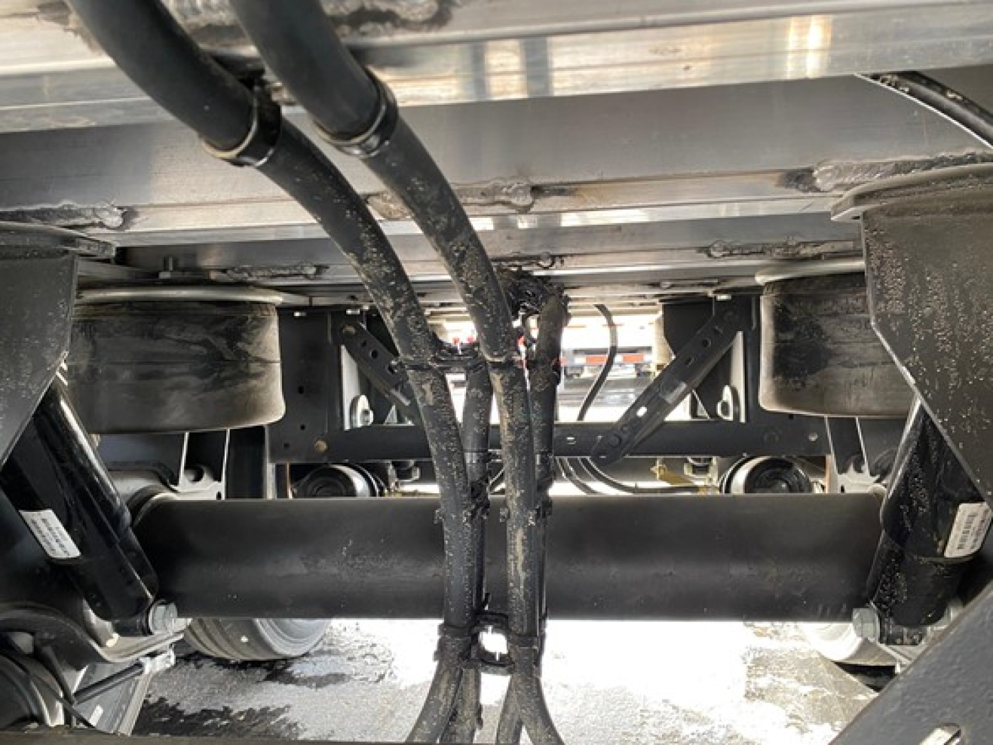 Underframe of a trailer showing suspension beams, axles, and hoses. Visible are black hydraulic lines, metal chassis, and wheels in the background.