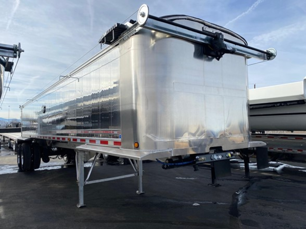 Dump trailer in silver metal with front hitch and landing gear, side view showing polished aluminum body and support legs, brand not visible.
