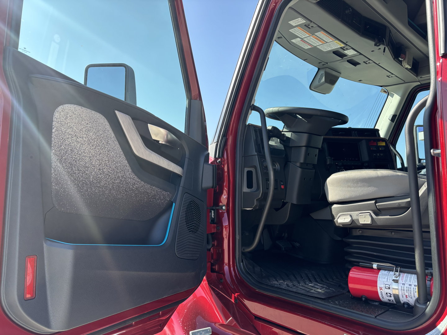 Cab interior of a 2027 Volvo VNL64T860 showing open driver door and dashboard, steering wheel, seat, and a fire extinguisher on the floor.
