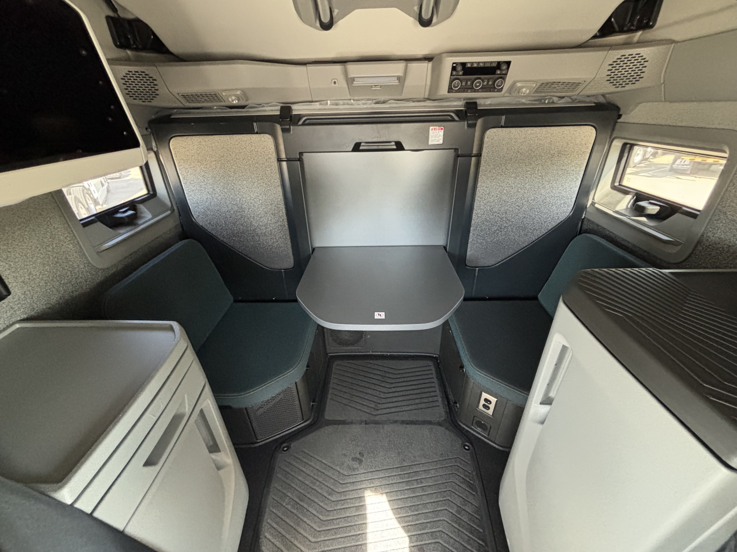 Cab interior of a 2027 Volvo VNL64T860 conventional sleeper showing compact dinette table with two curved seats and storage cabinets around, overhead controls, and grey/black color scheme.