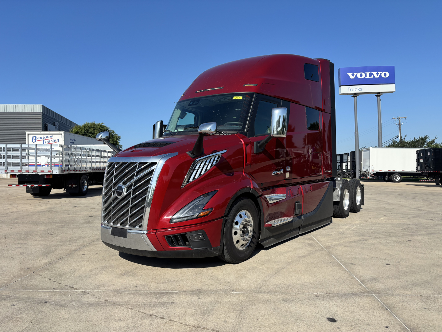 Volvo VNL64T860 truck in red seen from front-left angle with sleeper cab, chrome grille, and Volvo dealership sign in background.