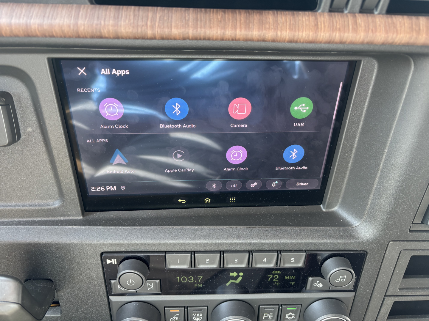 In-vehicle infotainment screen in a Volvo VNL64T860 showing apps and controls, with radio display below and wood trim around the dash.