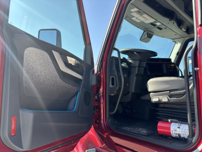 Cab interior of a 2027 Volvo VNL64T860 showing open driver door and dashboard, steering wheel, seat, and a fire extinguisher on the floor.