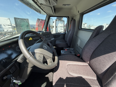 Truck cab interior of a 2026 Volvo VHD64B300 mixer showing steering wheel, dashboard, and driver seat with gear selector; visible window view of yard.