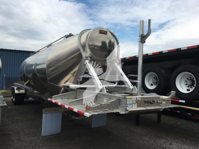 Circular Heil cementer/tank on trailer with support frame, rear view showing Heil logo on tank and chrome surface, with dual rear wheels and truck bed in background.