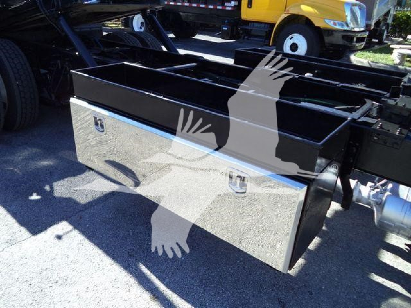 Truck bed toolbox mounted on chassis of a 2022 International MV rollback truck showing a large metallic toolbox with silver finish and black lid, side latches, and mounting points.