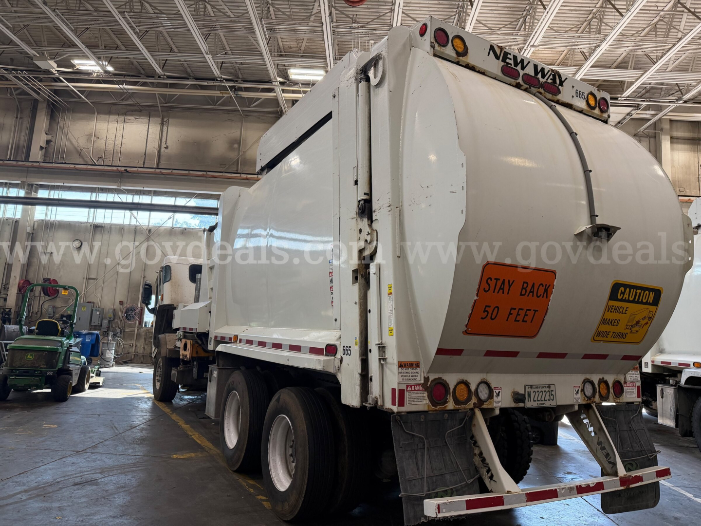 Rear and side view of a white Peterbilt garbage truck in a warehouse, with orange caution stickers reading STAY BACK 50 FEET and CAUTION; license plate area and rear lights visible.