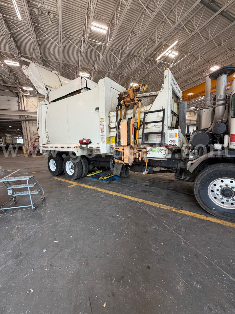 Peterbilt 520 truck in a maintenance bay with a large white refuse body mounted on a trailer; various labels and hydraulic components visible on the rear and side doors.